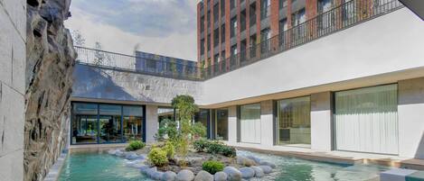 A modern hotel courtyard with a central pool, stone walls, and a multi-story building in the background.