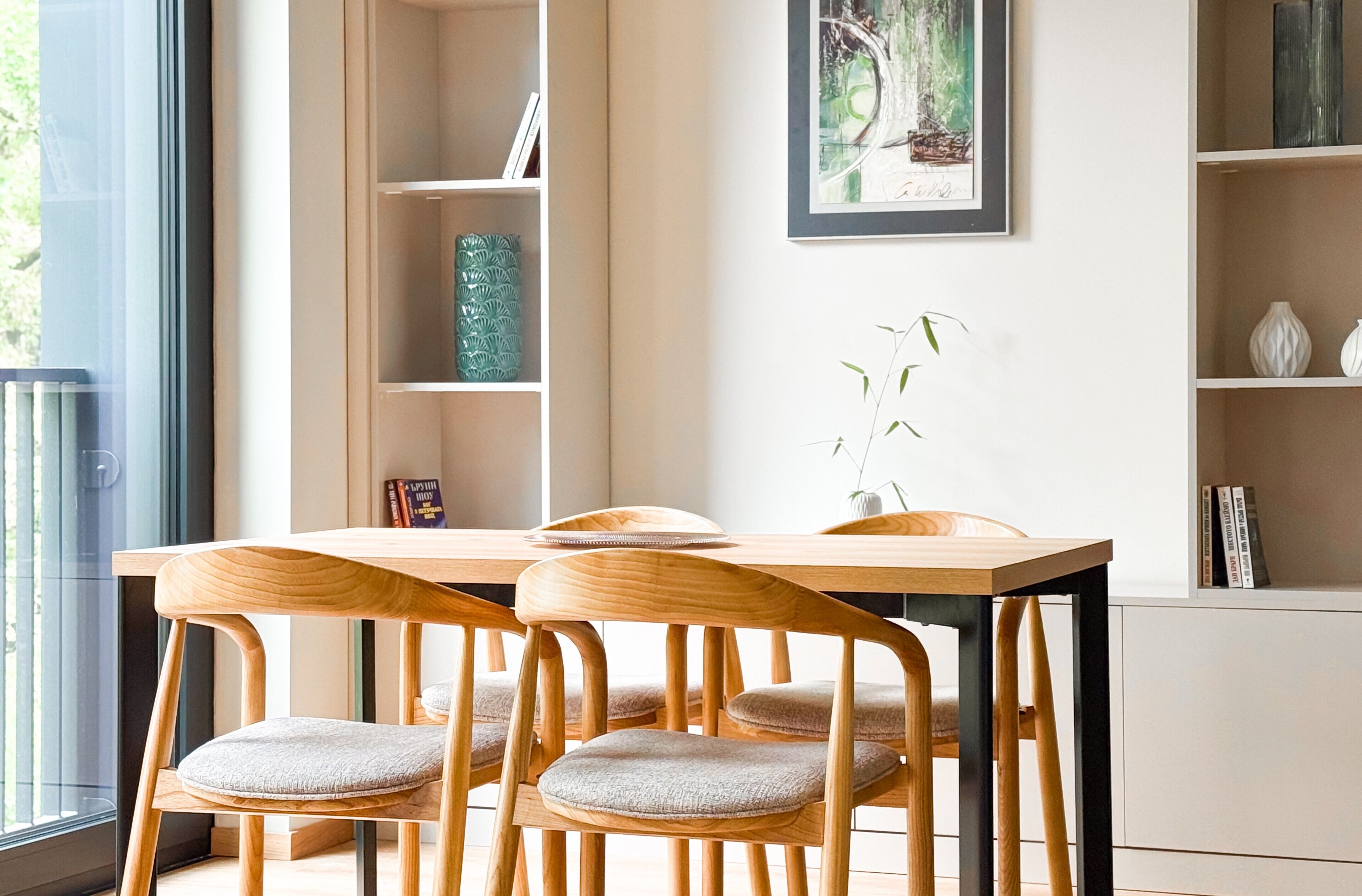 A modern dining area with a wooden table and chairs, a bookshelf, and a framed artwork on the wall.