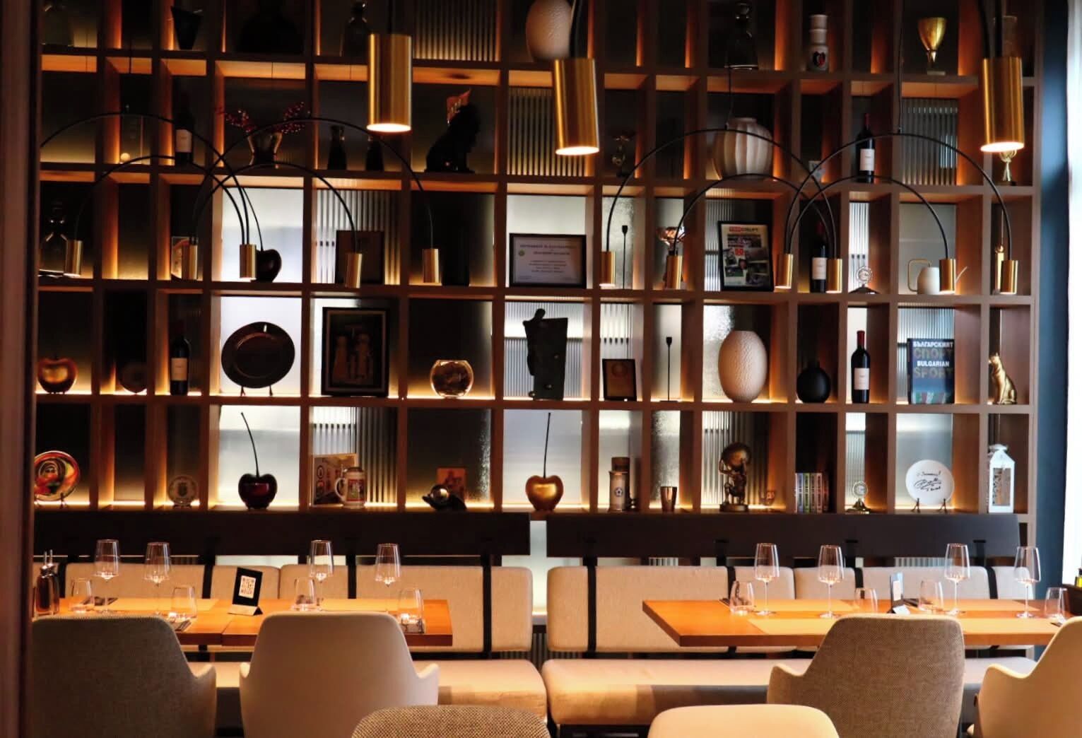 Un restaurante con un ambiente cálido, que presenta una pared de madera con estantes que exhiben varios objetos, y una zona de estar con mesas y sillas.