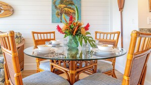 A dining area with a round glass table, wicker chairs, and a tropical-themed painting on the wall. - Makani A Kai A6 - On The Beach Steps To The Sand (Maalaea)