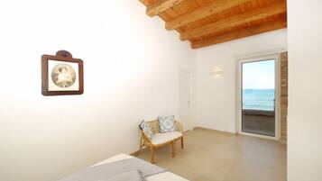 Habitación con techo de madera, pared blanca con un cuadro enmarcado, silla de mimbre con cojines, cama con manta gris y vista al mar a través de una puerta de cristal.