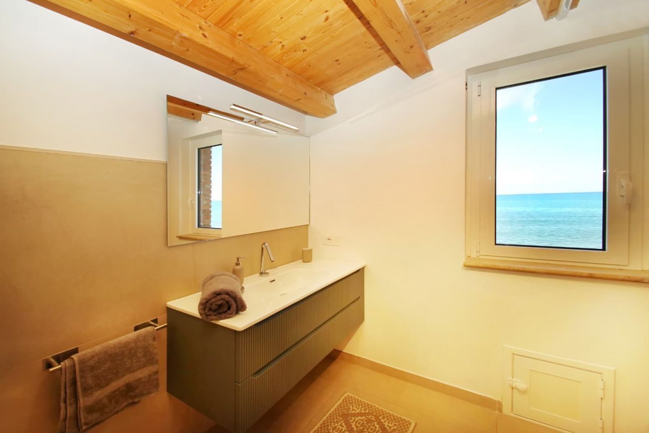A modern bathroom with a wooden ceiling, a large window with a view of the sea, a built-in sink, and a towel rack.