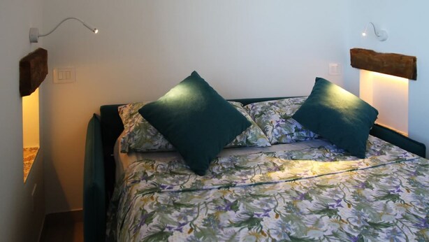 Un letto con biancheria da letto floreale e due cuscini verdi, un lampadario fissato al muro e una mensola in legno rustico.