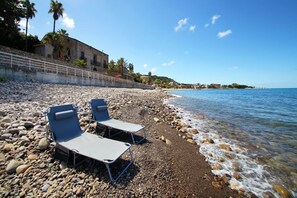 A pebble beach with two lounge chairs and a stone wall with a railing. - Relais Villa Giovanna -on the sea with garden (Marina di Caronia)
