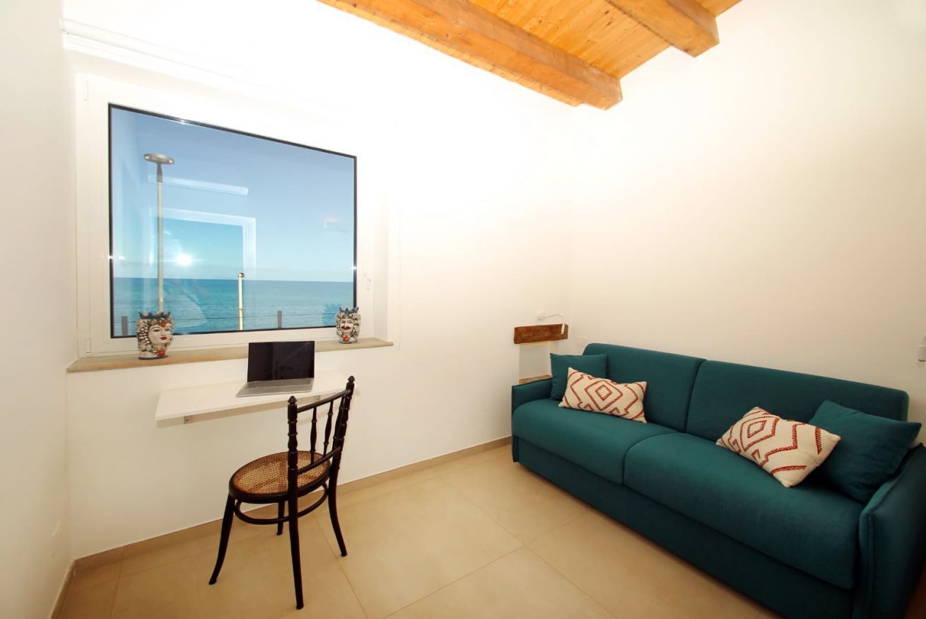 Ein Zimmer mit einem großen Fenster mit Meerblick, ein türkisfarbenes Sofa mit gemusterten Kissen, ein Holzstuhl und ein kleiner Tisch mit einem Laptop.