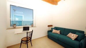 Una stanza con una grande finestra vista mare, un divano color petrolio con cuscini decorati, una sedia in legno e un tavolino con un portatile.
