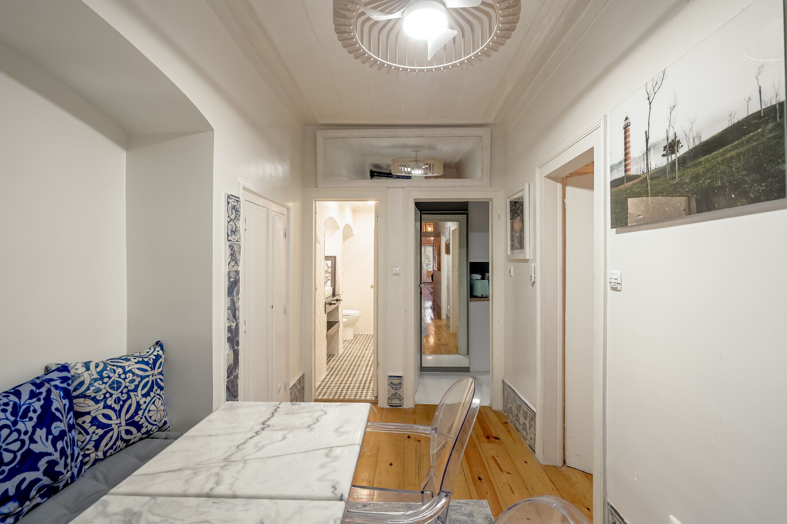 Un pasillo estrecho con una cama, un sillón transparente y un refrigerador.