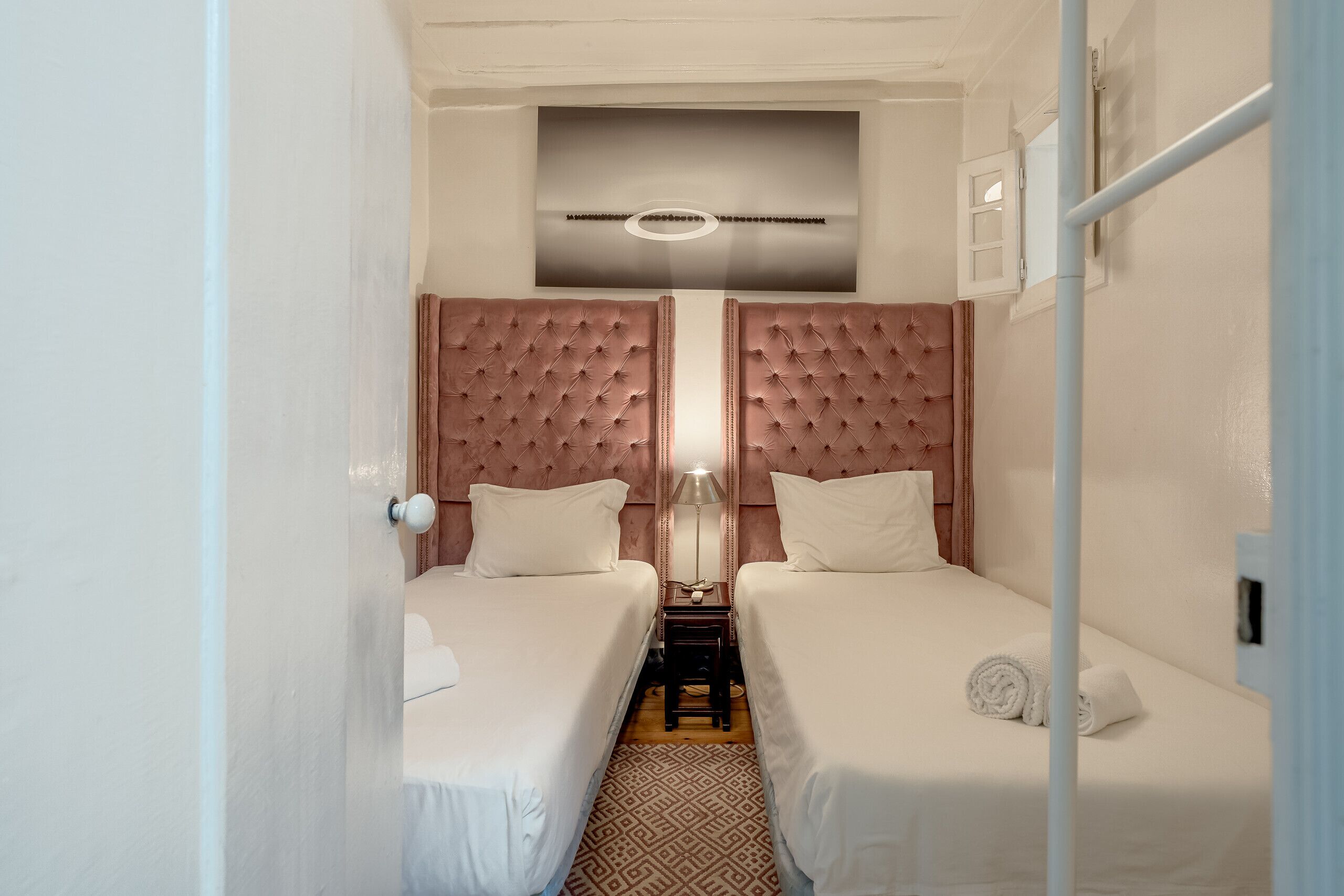 A hotel room with two beds, a patterned rug, a ceiling light, and a small table with a lamp.