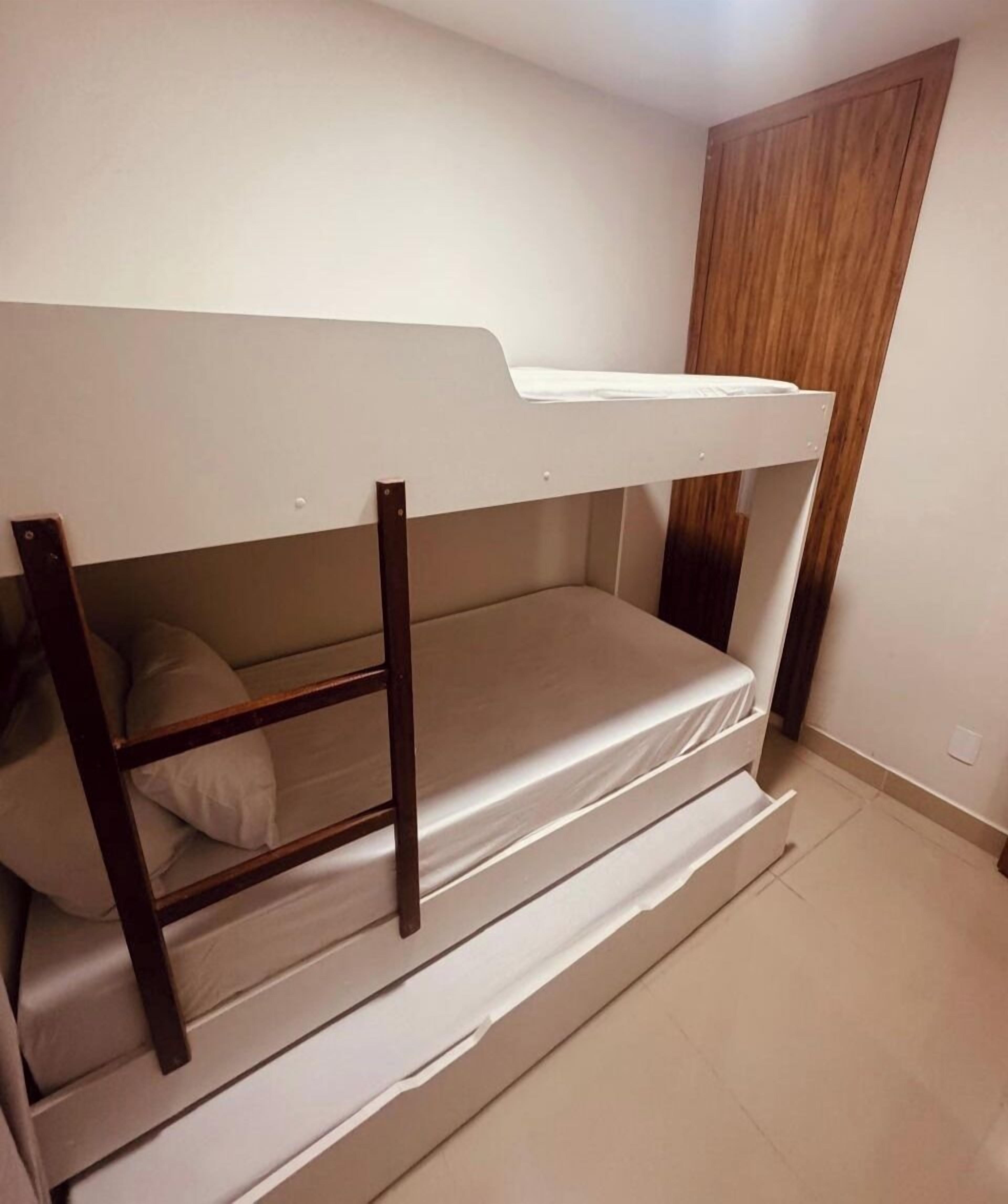 Una cama litera con estructura de madera y ropa de cama blanca en una habitación con suelo de baldosas y puerta de madera.