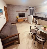 A compact living space with a sofa, a TV, a dining table with chairs, a kitchen area, and a small balcony.
