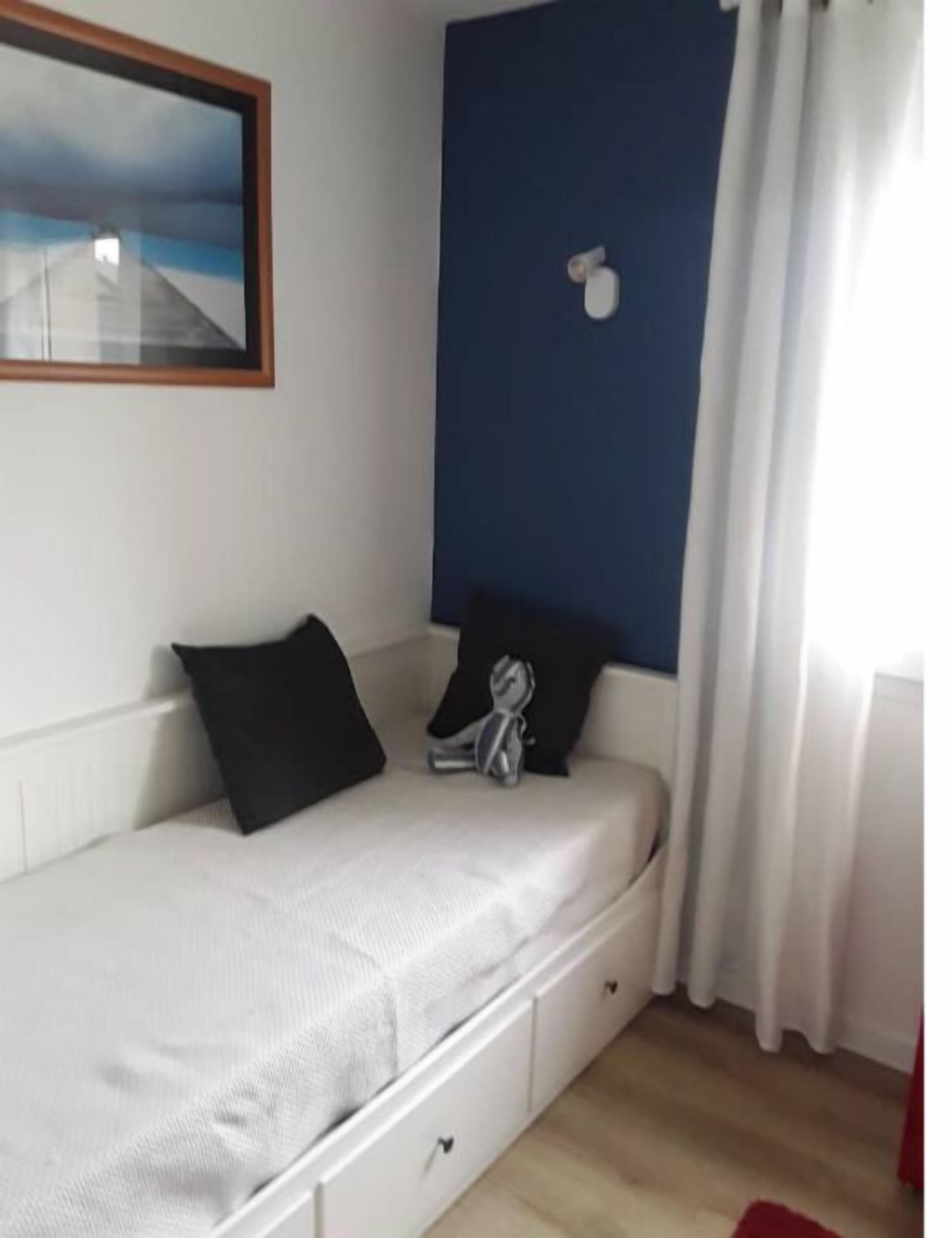 A single-bed room with a white bed, black pillows, a small bedside table, a nightstand, a blue accent wall, a framed picture, and a window with white curtains.