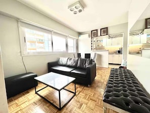 A modern living room with a leather sofa, a glass-top coffee table, and a black tufted bench. A kitchen area is visible in the background with white cabinets and a microwave. - -Belgrano- (Buenos Aires)