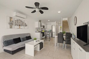 Un salon moderne avec un canapé gris, une table basse blanche et un coin repas équipé d’un téléviseur et de chaises.