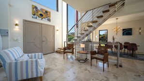 Ampla sala de estar com sofá listrado, mesa de jantar com cadeiras e escada com corrimão.
