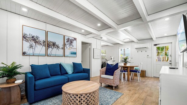 A modern living room with a blue sofa, a round wooden coffee table, and a wicker chair. There are three framed pictures on the wall, a flat-screen TV mounted on the wall, and a dining area with a table and chairs. - A Charming Beach Cottage - Unit B (Siesta Key)