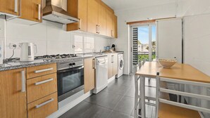 A modern kitchen with wooden cabinets, a gas stove, a washing machine, and a dining area with a table and chairs. - Apartment 'Campo Mar - T2 Com Piscina' with Shared Pool, Wi-Fi and Air Conditioning (São Martinho do Porto)