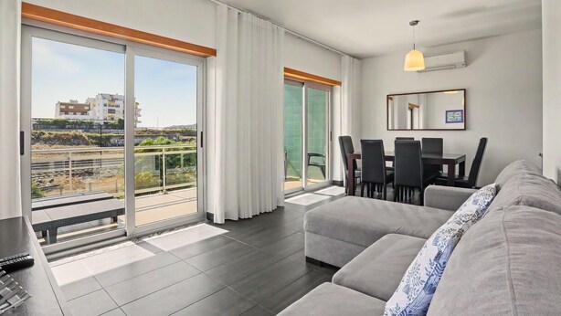 A modern living room with a sofa, dining table, chairs, and a balcony with a view. - Apartment 'Campo Mar - T2 Com Piscina' with Shared Pool, Wi-Fi and Air Conditioning (São Martinho do Porto)