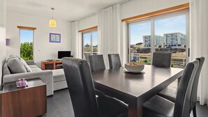 A modern hotel room with a dining area, large windows, and a view of buildings. - Apartment 'Campo Mar - T2 Com Piscina' with Shared Pool, Wi-Fi and Air Conditioning (São Martinho do Porto)