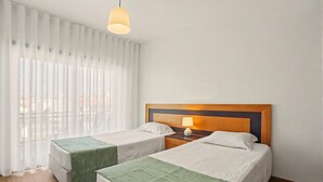 A hotel room with two beds, a wooden headboard, a nightstand with a lamp, and sheer curtains. - Apartment 'Campo Mar - T2 Com Piscina' with Shared Pool, Wi-Fi and Air Conditioning (São Martinho do Porto)