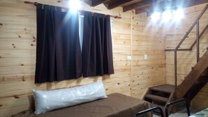 Quarto de cabana de madeira com cama, janela com cortinas escuras, um banquinho pequeno e escada de madeira.