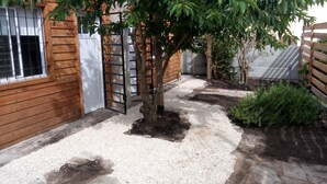 Uma área de jardim com uma árvore, caminho de cascalho, cerca de madeira e um edifício com janela.