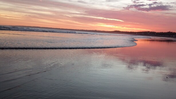 Uma praia com areia molhada e ondas suaves, céu claro e um pôr do sol.