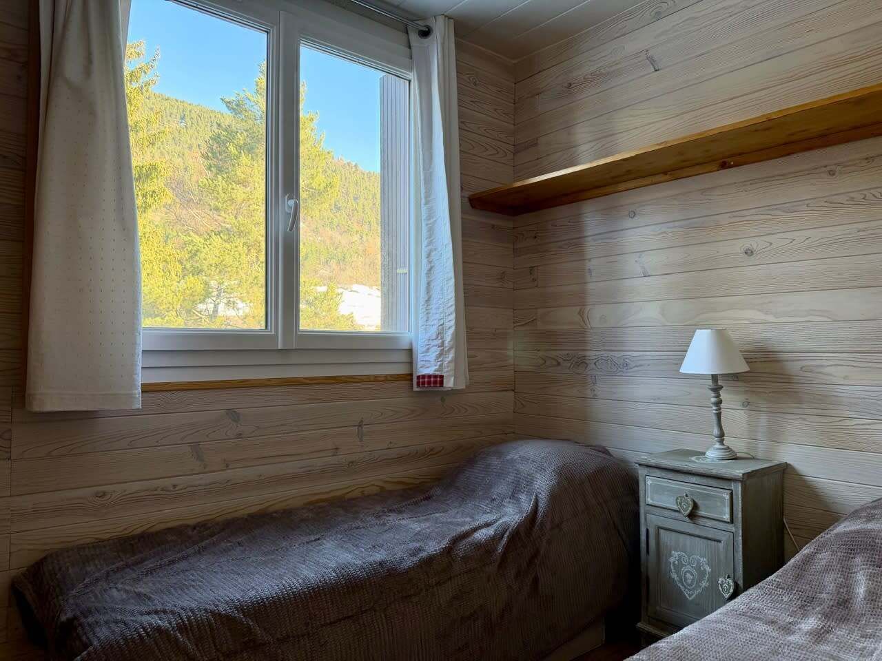 Ein kleiner, gemütlicher Raum mit Holzwänden, einem Einzelbett, einem Nachttisch mit Lampe und einem Fenster mit Vorhängen.