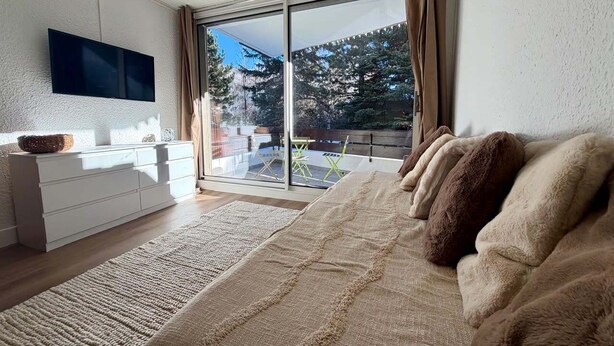 A modern bedroom with a large bed, a flat-screen TV, a white dresser, and a sliding glass door leading to a balcony with outdoor furniture. - Pleasant stay in La Salle-les-Alpes for 4 people (La Salle-les-Alpes)
