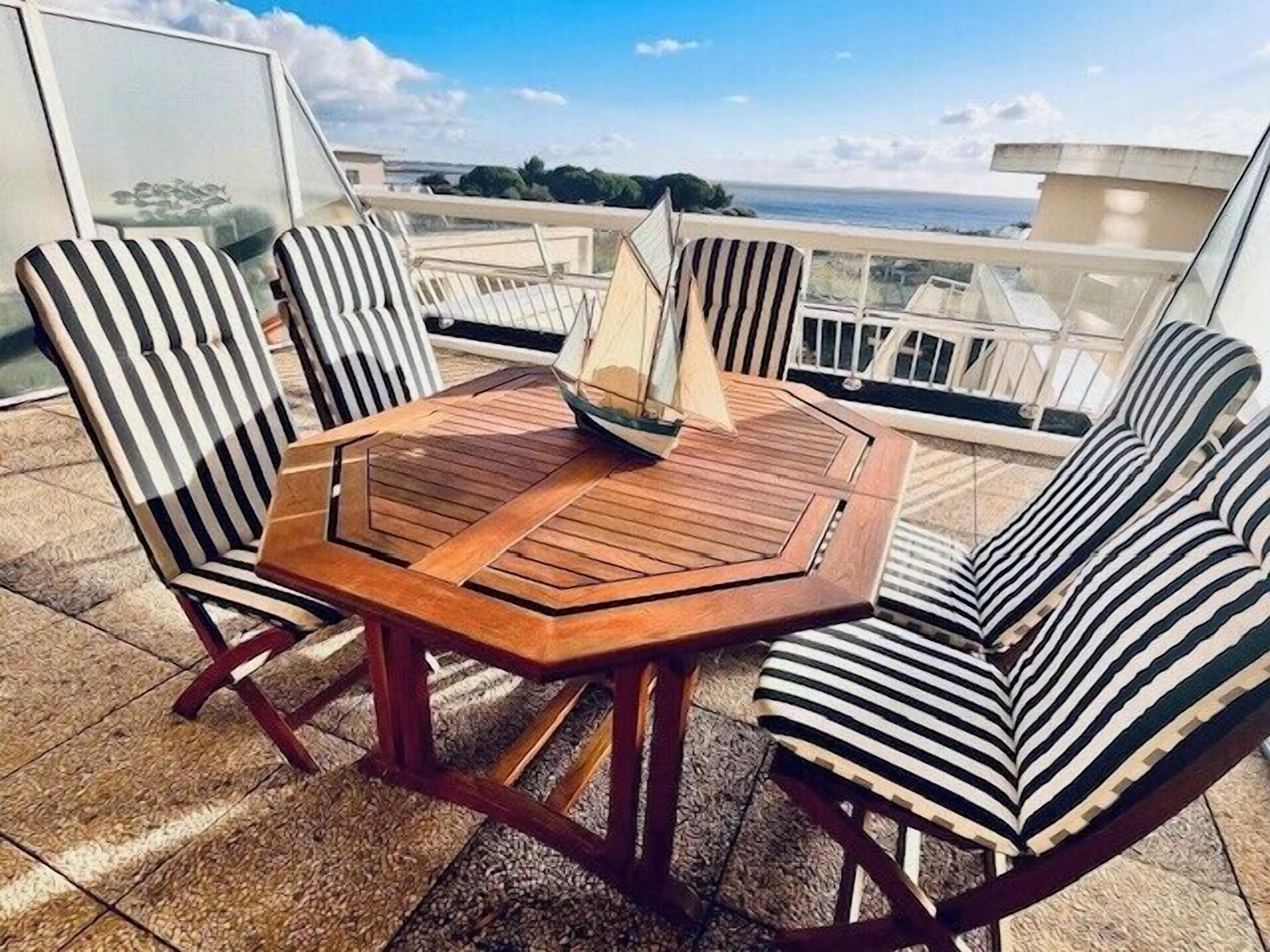 A wooden table with six striped chairs on a balcony with a view of the sea.