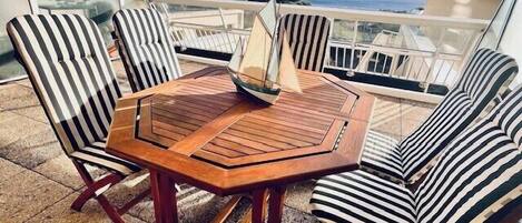 A wooden table with six striped chairs on a balcony with a view of the sea.