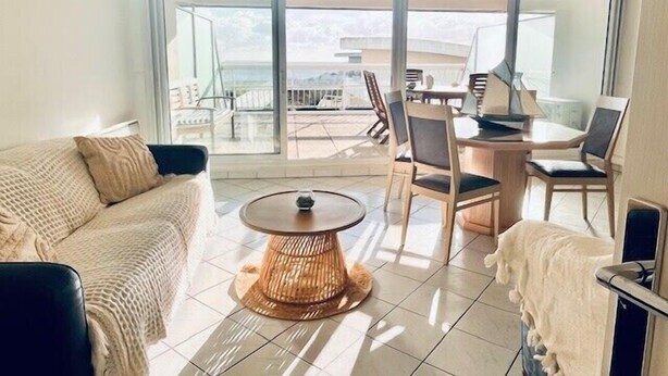 Un soggiorno moderno con un divano, un tavolino rotondo e sedie, ampie finestre che si affacciano su un balcone e sull'esterno.