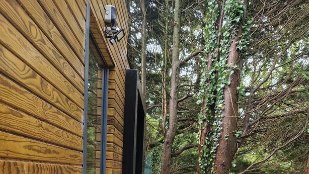 Une terrasse en bois avec deux chaises bleues et une rambarde en verre, entourée d’arbres.