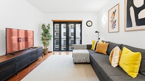 Un salon moderne avec un canapé gris, des coussins jaunes, un téléviseur à écran plat, une plante en pot et une grande fenêtre donnant sur des immeubles.