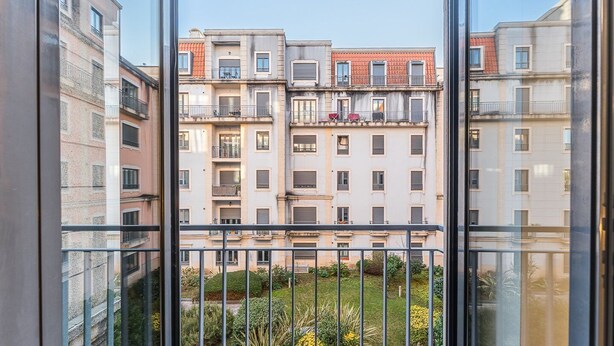 Une vue depuis un balcon donnant sur une rangée d’immeubles à plusieurs étages, chacun doté de balcons, ainsi que sur un jardin planté de diverses espèces et un ciel dégagé.