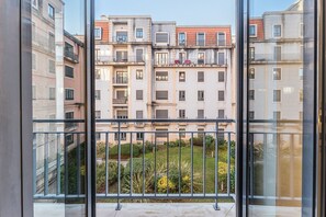 Vista de uma varanda com vista para uma fileira de edifícios com várias varandas, uma área de jardim com diversas plantas e um céu limpo.