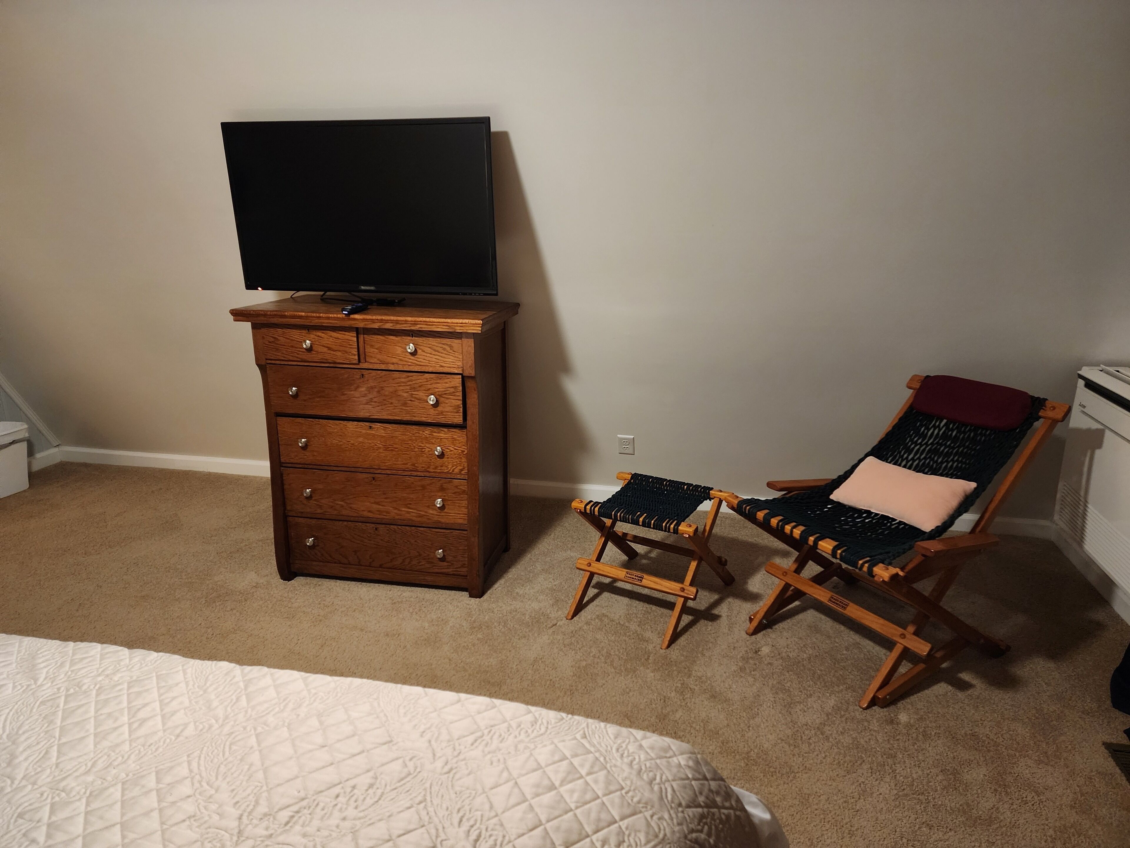 A bedroom with a wooden dresser, a television on top, a bed with a quilted cover, and two wicker chairs with cushions.