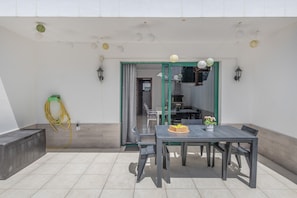 Une terrasse extérieure avec une table et des chaises, une porte à cadre vert et un tuyau d’arrosage fixé au mur.