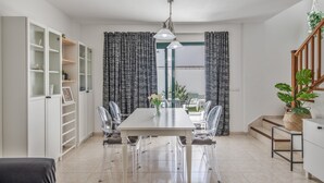 Un comedor moderno con una mesa blanca, sillas transparentes, una lámpara colgante y una escalera con una planta.