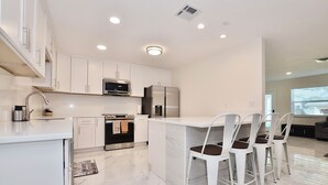 Una cocina moderna con armarios blancos, una isla central con taburetes de bar, electrodomésticos de acero inoxidable y suelo de mármol.