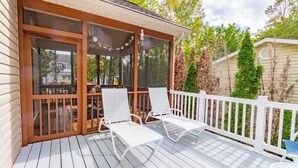 En overdækket terrasse med hvide liggestole og et bord, omgivet af et hvidt hegn og frodig vegetation.