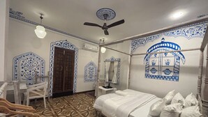 Een kamer met een bed, een bureau en een stoel. Er is een plafondventilator en een raam met decoratieve details.