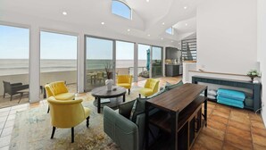 A spacious living area with a large window overlooking the sea, a wooden dining table, yellow armchairs, and a sofa.