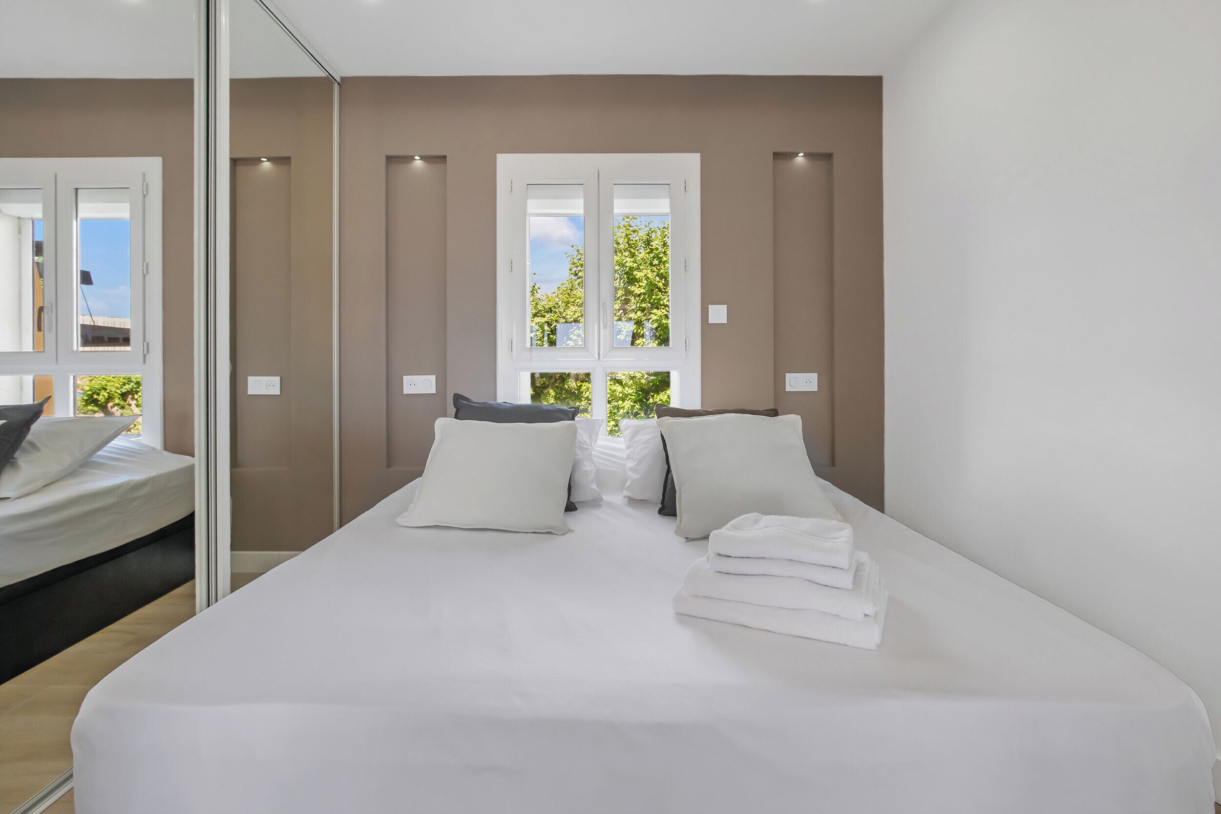 A modern hotel room with a large bed, a mirror, and a view of greenery through the window.