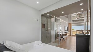 Moderni hotellihuone, jossa on sänky, lasiseinäinen suihkutila, ruokailualue ja näkymä ulos.