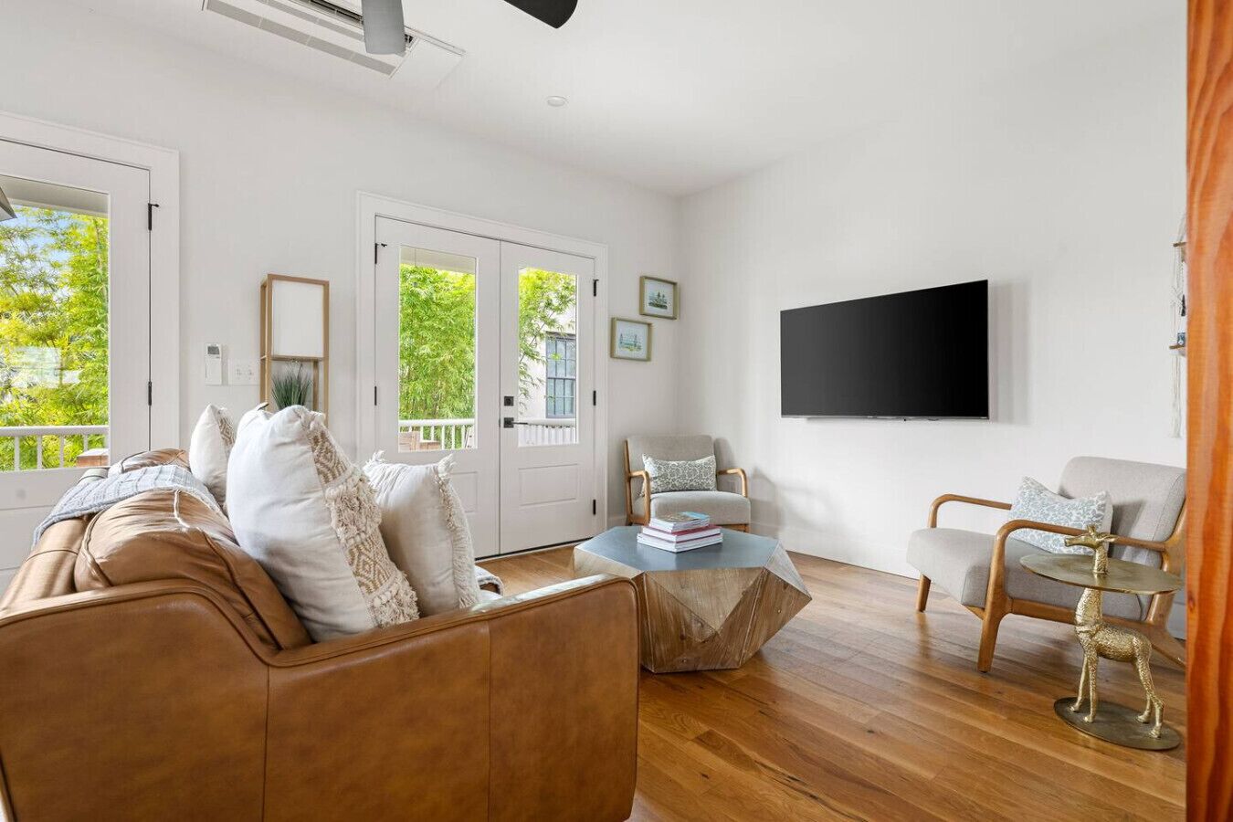 Amplia sala de estar con un sofá de cuero marrón, una mesa de centro de madera y dos sillones. Hay un televisor de pantalla plana grande montado en la pared, y una puerta que da a un balcón con vistas a zonas verdes.