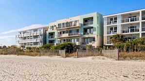 Un hôtel en bord de mer doté de nombreux balcons, une plage de sable en premier plan et un ciel bleu dégagé.