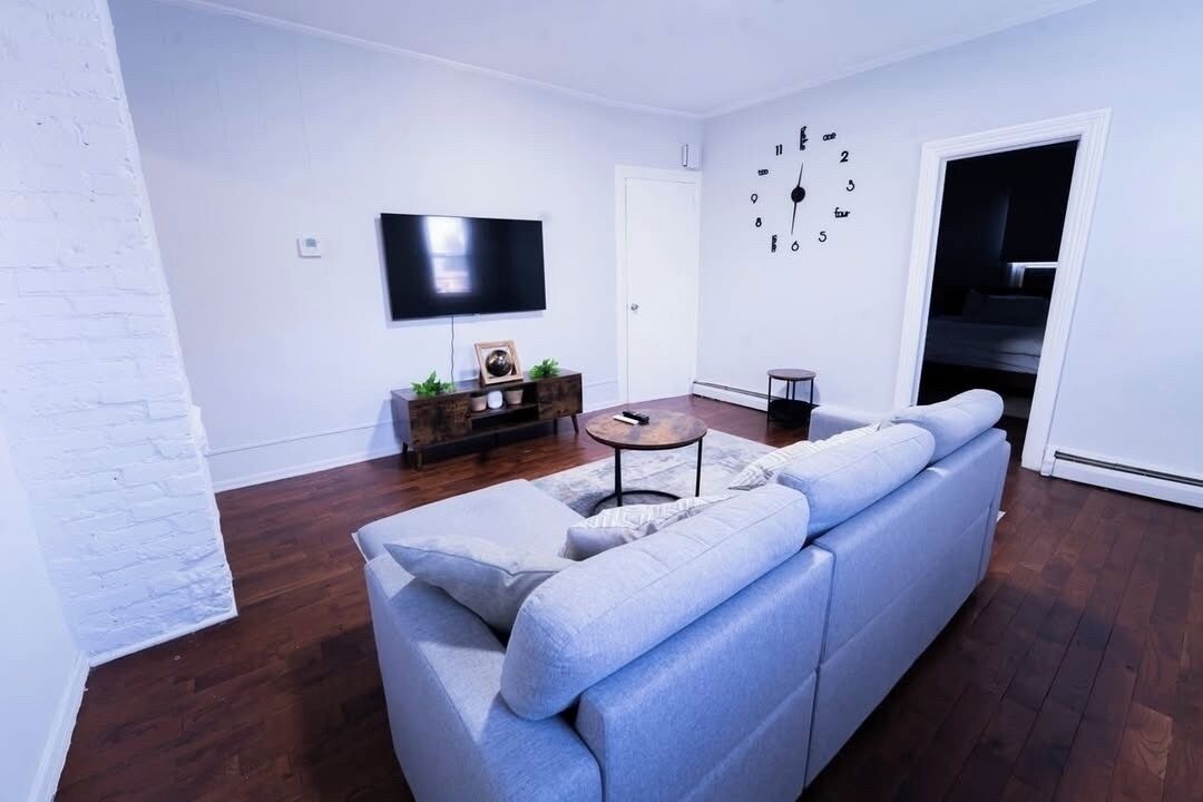 Un salon moderne avec un canapé d’angle, un téléviseur à écran plat, une petite table ronde et une horloge murale décorative.