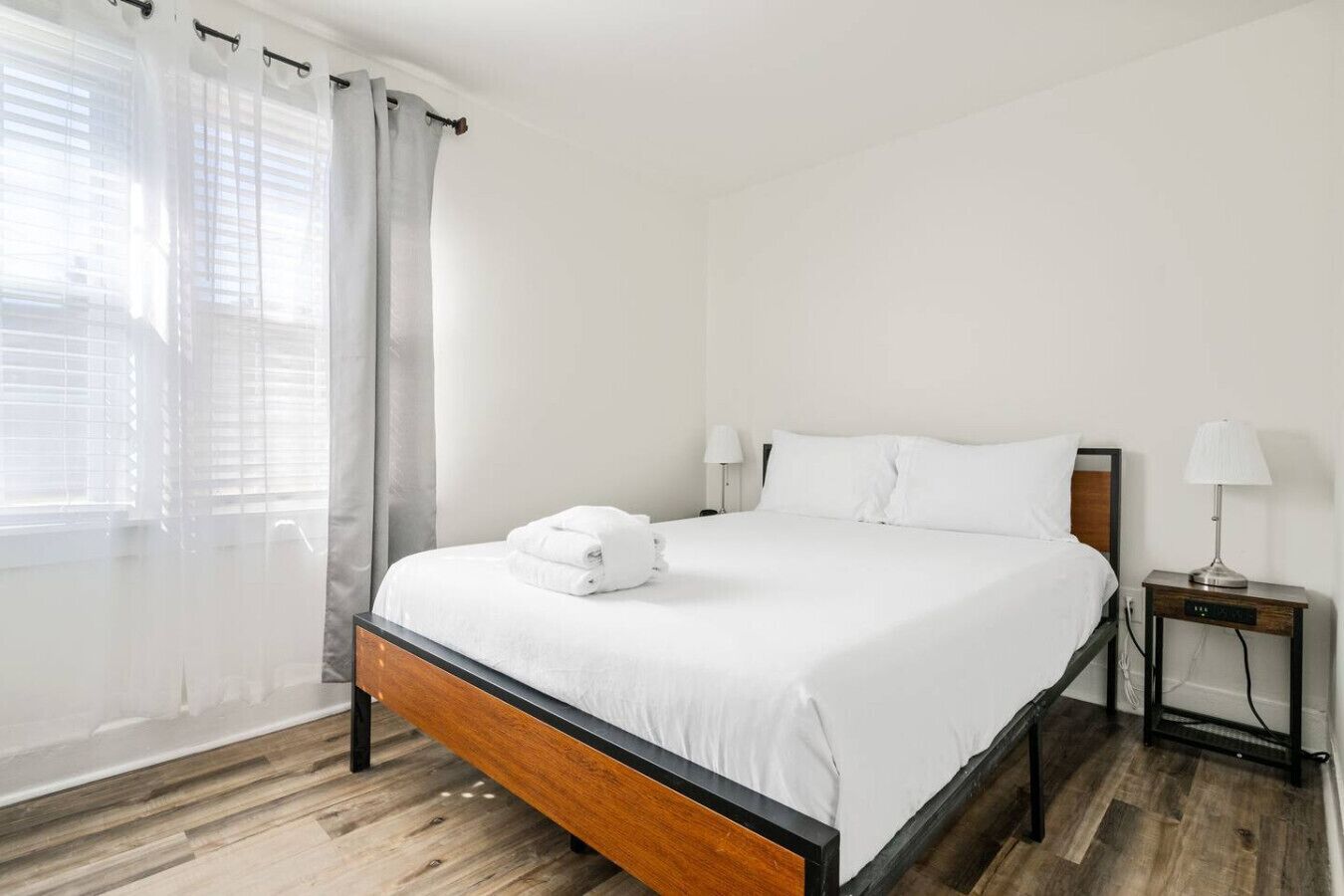A neatly made bed with white linens and a folded towel, a bedside table with a lamp, and a window with sheer curtains.