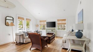 A spacious living room with a high ceiling, wooden flooring, and a large window with a view of greenery. There is a television on a wooden stand, a wicker chair, and a white sofa. - Beach Maverick - 3 BR 2 Bath 1 Block to Center St! (Folly Beach)