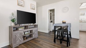 Ein Wohnzimmer mit Flachbildfernseher auf einem Holz-TV-Möbel, eine Essecke mit Tisch und Stühlen sowie eine durch einen offenen Durchgang sichtbare Küche.